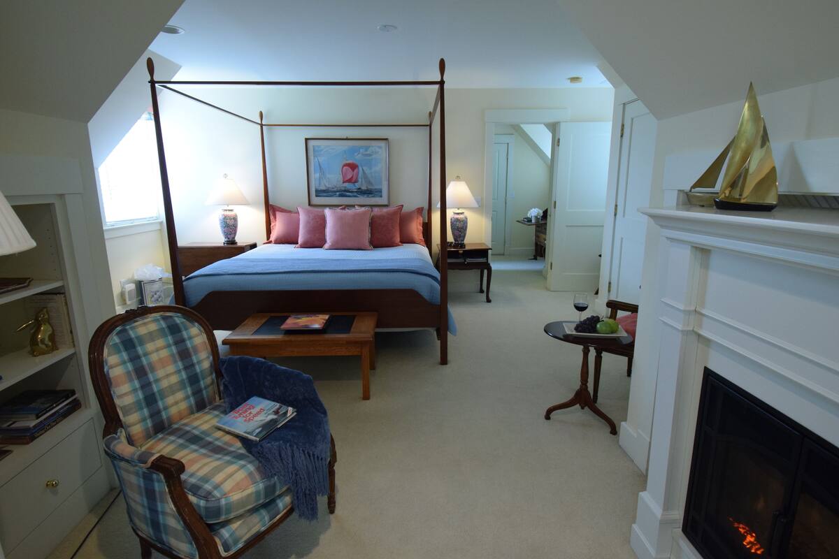 A bedroom with a four-poster bed, a fireplace, a chair, a bedside table, and a painting on the wall.