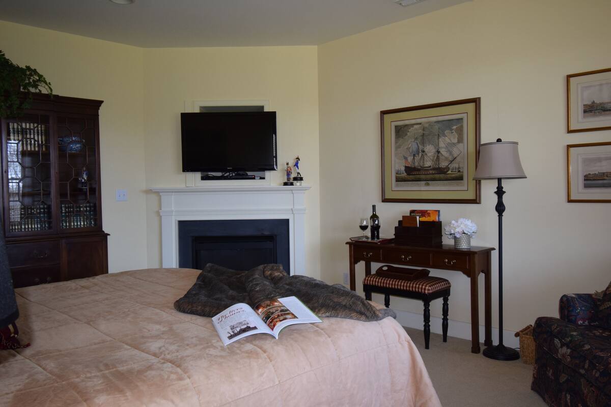 A bedroom with a large bed, a television above a fireplace, a desk with a lamp, and framed pictures on the wall.