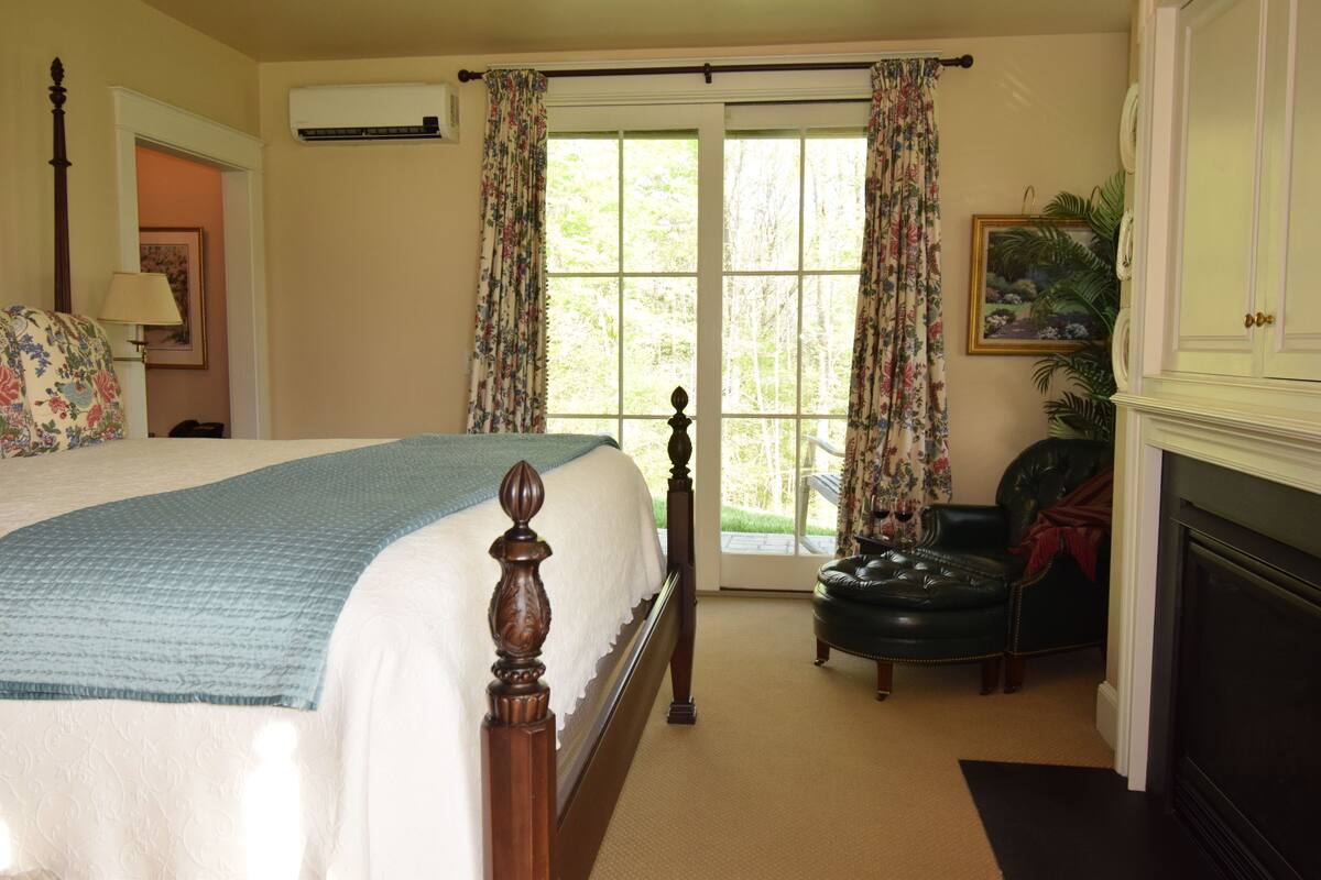 A bedroom with a large bed, a fireplace, a chair, and a door leading to a balcony.