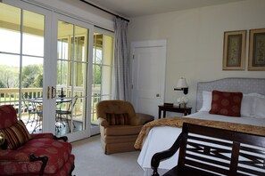 A hotel room with a large sliding glass door leading to a balcony, a bed with a patterned bedspread, a red armchair, a wooden chair, and a small bedside table with a lamp.