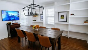 A dining area with a wooden table, six chairs, a decorative centerpiece, a television on the wall, and white shelving with books and framed pictures. - Field House | Game Room | DOWNTOWN Dells | Basketball Court| Arcade (Wisconsin Dells)