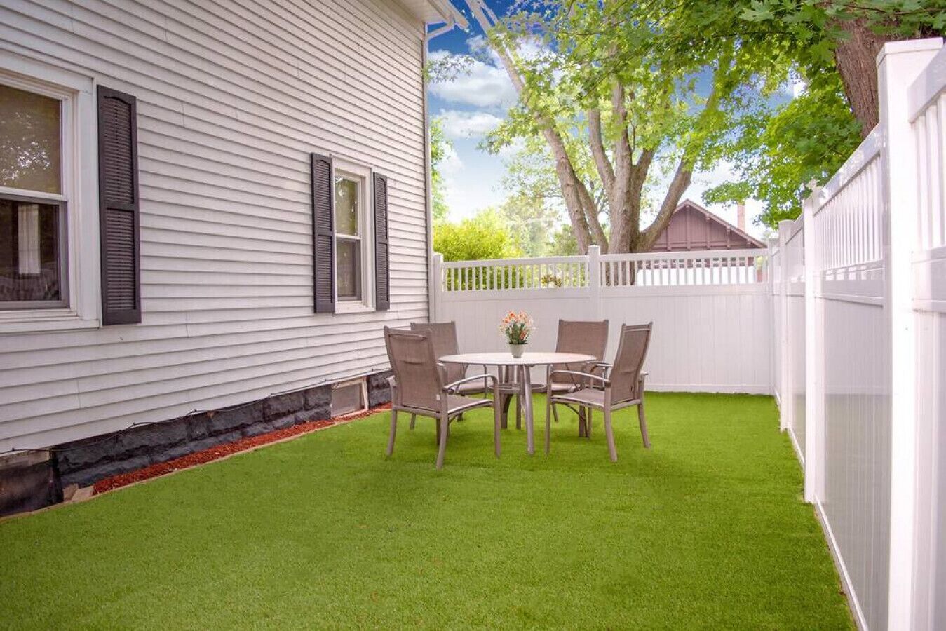 Un jardin avec une clôture blanche, une table et des chaises, et un arbre.