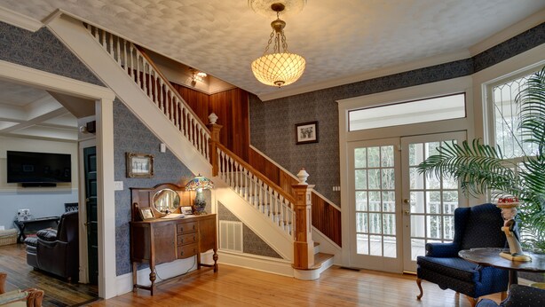 A spacious living room with hardwood floors, a wooden staircase, a chandelier, and a large window with a view. - Historic Hot Springs Home < 1 Mi to Oaklawn! (Hot Springs)