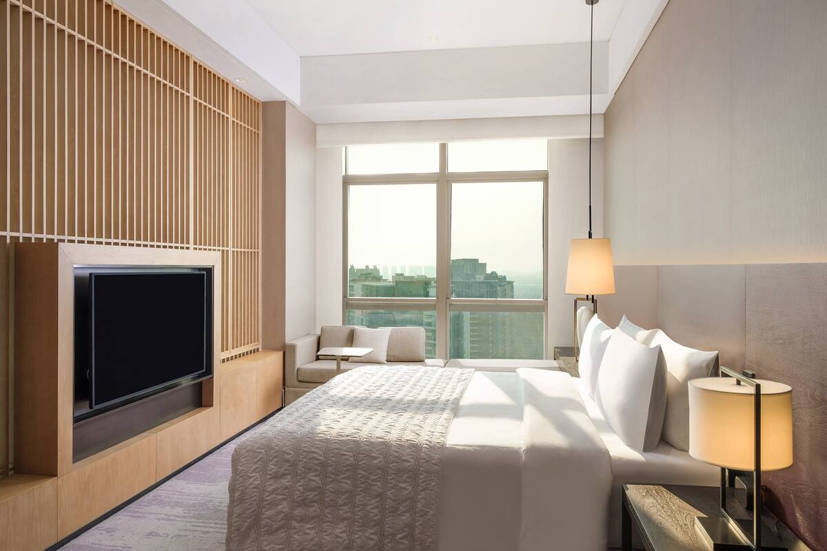 Una habitación de hotel moderna con una cama grande, un televisor de pantalla plana, un área de descanso con una mesita y un ventanal con vistas a la ciudad.
