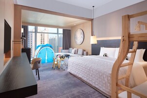 Habitación de hotel con cama, escritorio, silla, una mesita pequeña y un ventanal con vistas a edificios.