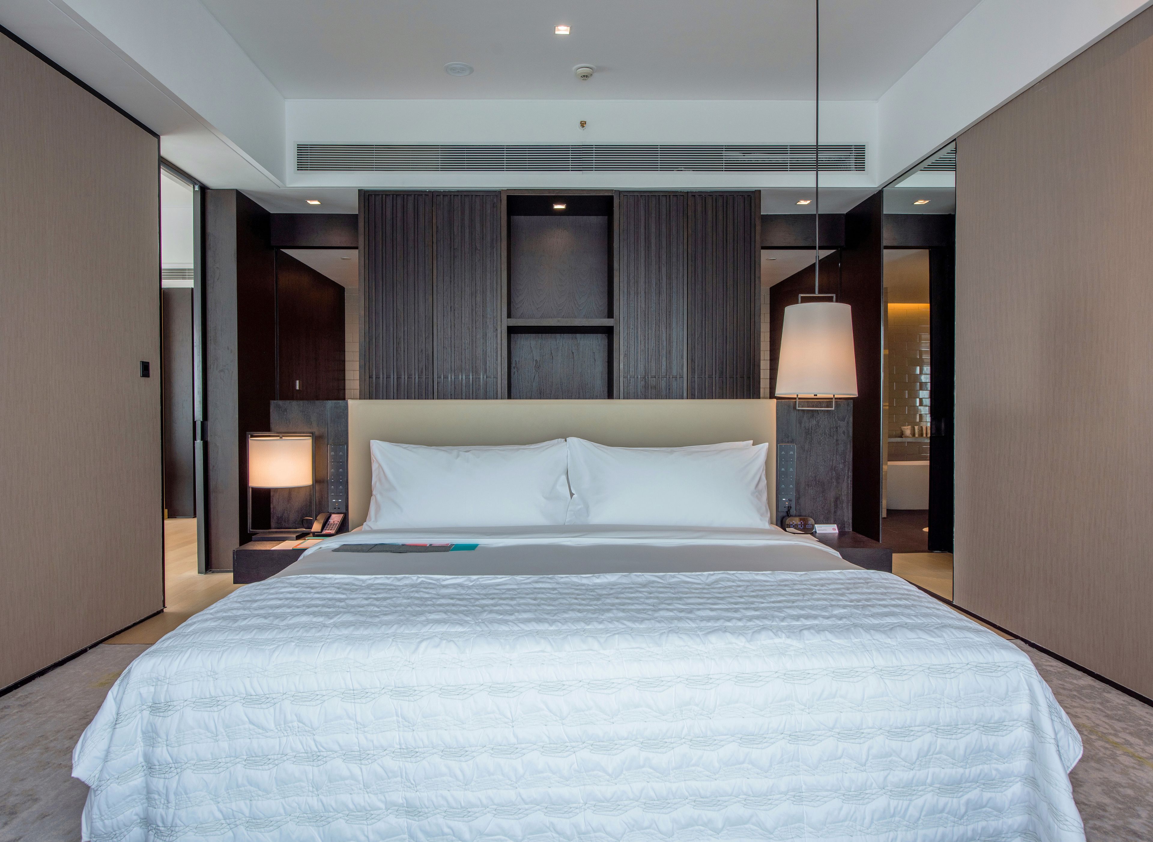 A modern hotel room with a large bed, a bedside table with a lamp, and a large mirror.