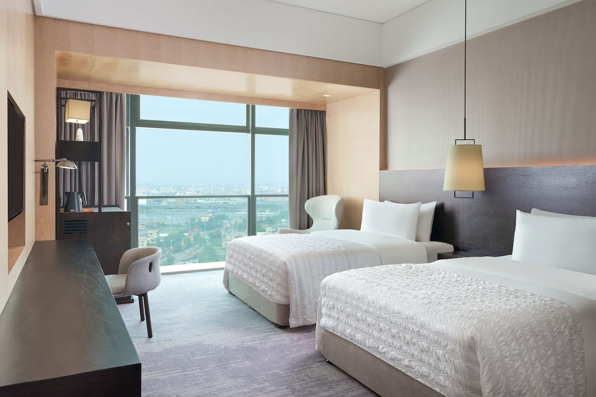 Habitación de hotel con dos camas, un escritorio, una silla y un ventanal con vistas a la ciudad.