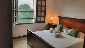 A hotel room with a bed, a window with a view of greenery, a bedside table with a lamp, and an air conditioning unit.