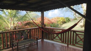 A covered porch with a hammock, wooden bench, and a view of a garden with palm trees and flowering plants. - Top of the Hill Two Bedroom Apartment, Rodney Bay (Gros Islet)