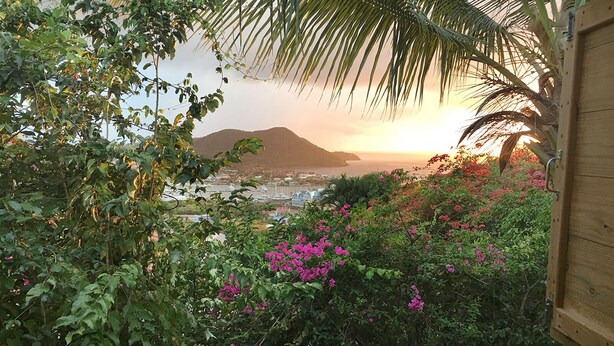 A view from a wooden structure overlooking a coastal area with mountains, lush greenery, and pink flowers. - Top of the Hill Two Bedroom Apartment, Rodney Bay (Gros Islet)