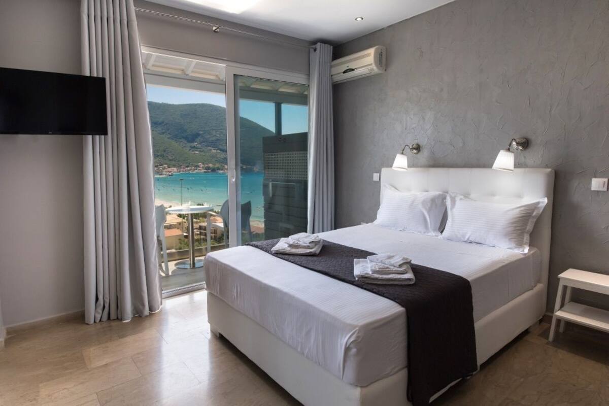 Habitación de hotel moderna con una cama grande, un televisor montado en la pared y un balcón con vistas al mar y las colinas.