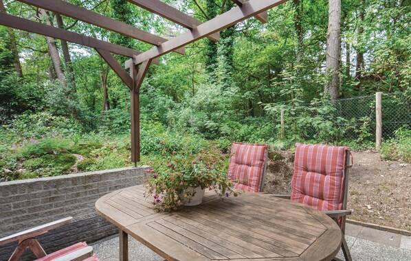 Una mesa de madera con una planta en maceta, dos cojines a rayas y un banco bajo una pérgola de madera en una zona boscosa.