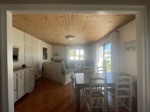 Une pièce avec un plafond en bois, une table à manger avec des chaises, un coin cuisine avec des placards et une fenêtre avec des rideaux.