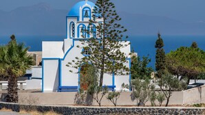 Un edificio blanco con detalles azules y un campanario, rodeado de árboles y un muro de piedra, con vista a un cuerpo de agua.