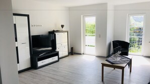 A modern living room with a flat-screen TV, a black sofa, a coffee table, and a sliding door leading to a balcony with a view of greenery and buildings. - Appartemens Haus Maria Anna (Bad Ems)