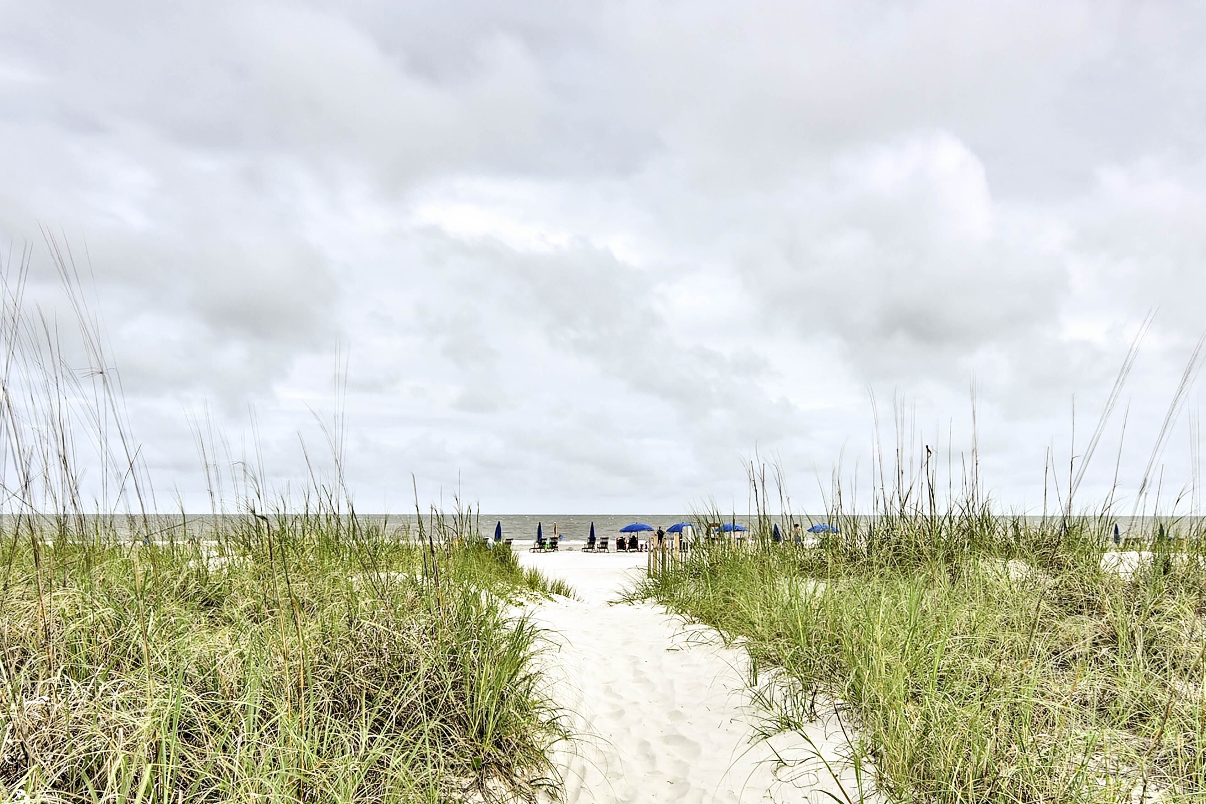 A sandy beach path flanked by tall grass leading to a pier with umbrellas.