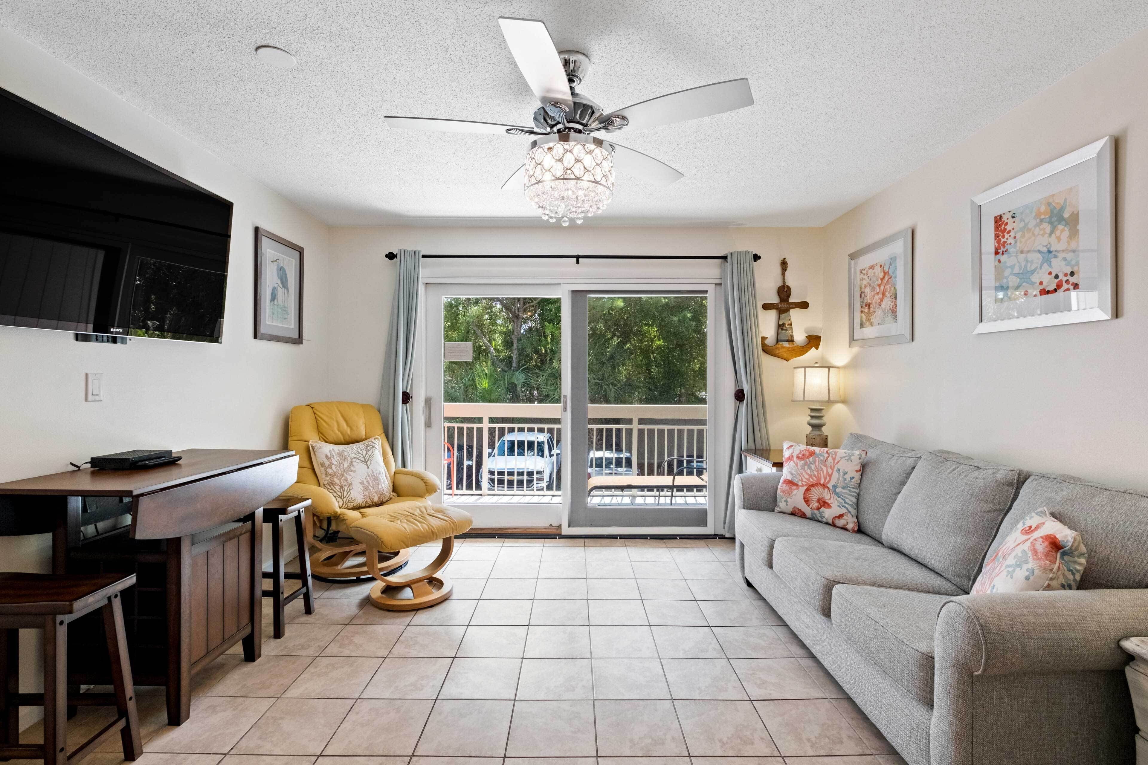 A living room with a sofa, a dining table, a yellow chair, a ceiling fan, and a view of the outdoors through a sliding glass door.