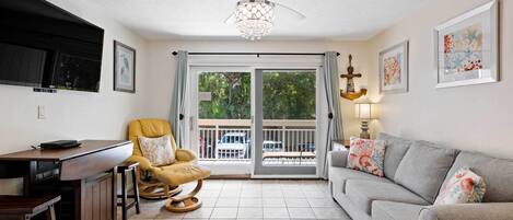 A living room with a sofa, a dining table, a yellow chair, a ceiling fan, and a view of the outdoors through a sliding glass door.