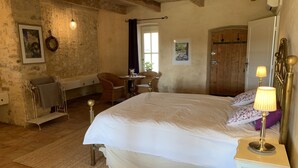 Un dormitorio con una cama grande, una mesita de noche con lámpara, una silla, una pequeña mesa con un jarrón y una puerta de madera.