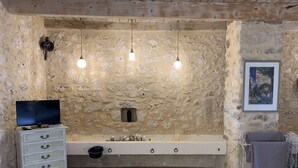 Una habitación rústica con pared de piedra, un banco blanco, un televisor en una cómoda y un cuadro en la pared.