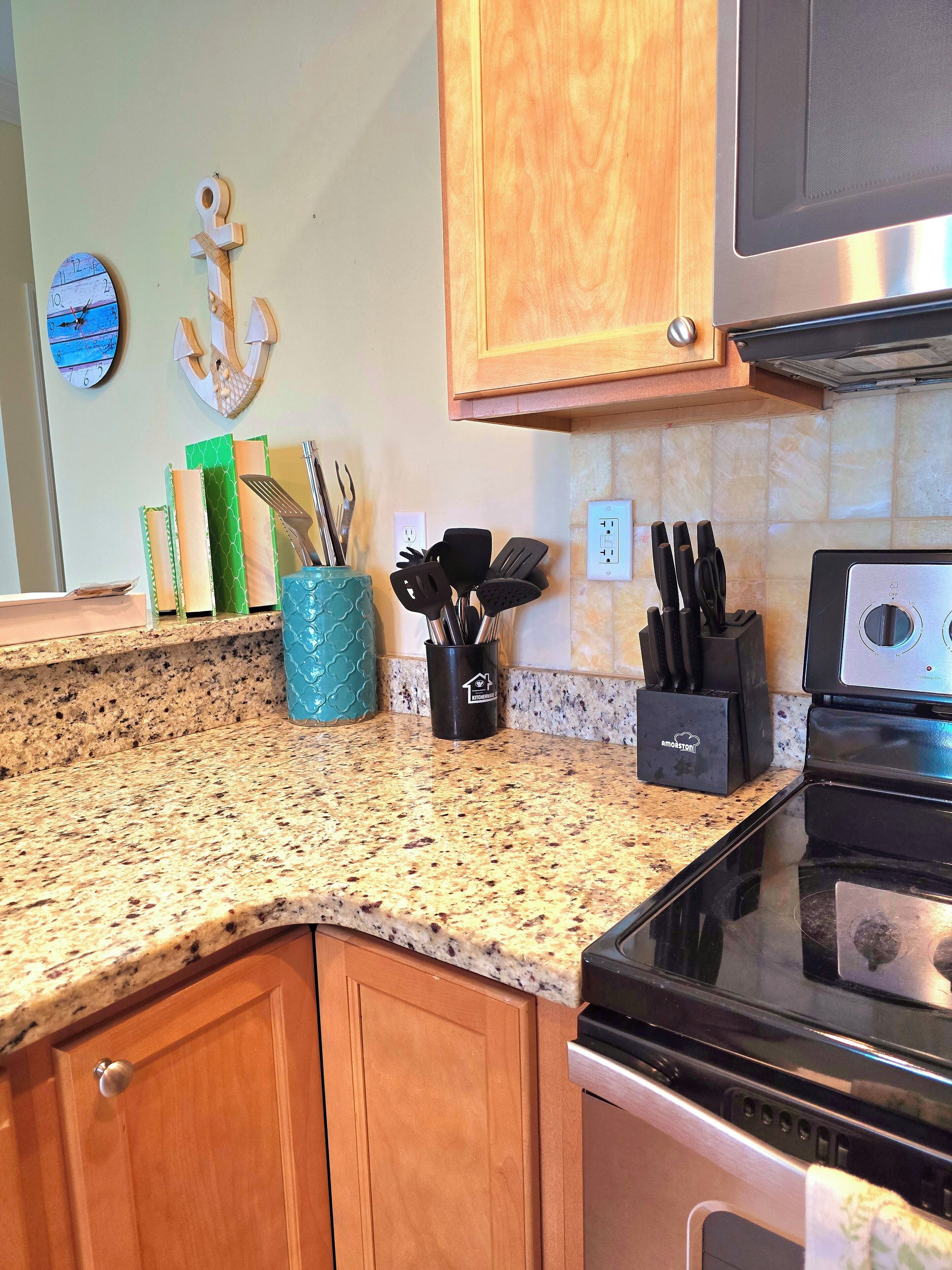 A kitchen with granite countertops, wooden cabinets, a stainless steel oven, and a microwave. There are various kitchen utensils and a decorative anchor on the wall.
