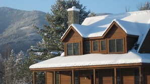 Une cabane en bois, dont le toit est recouvert de neige, avec de grandes fenêtres et une cheminée en pierre.