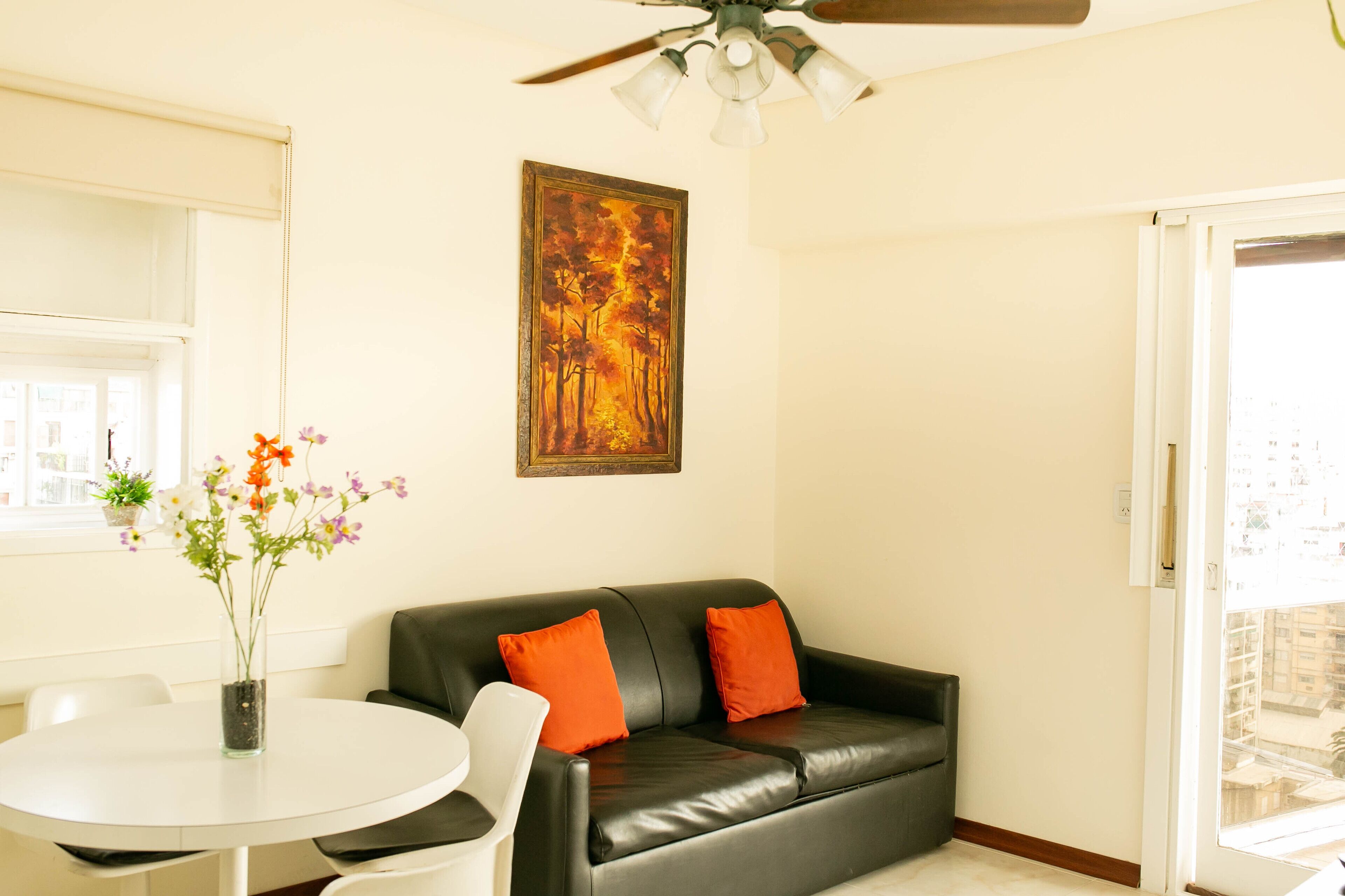 A living room with a black leather sofa, two orange cushions, a white table, and a vase with flowers.