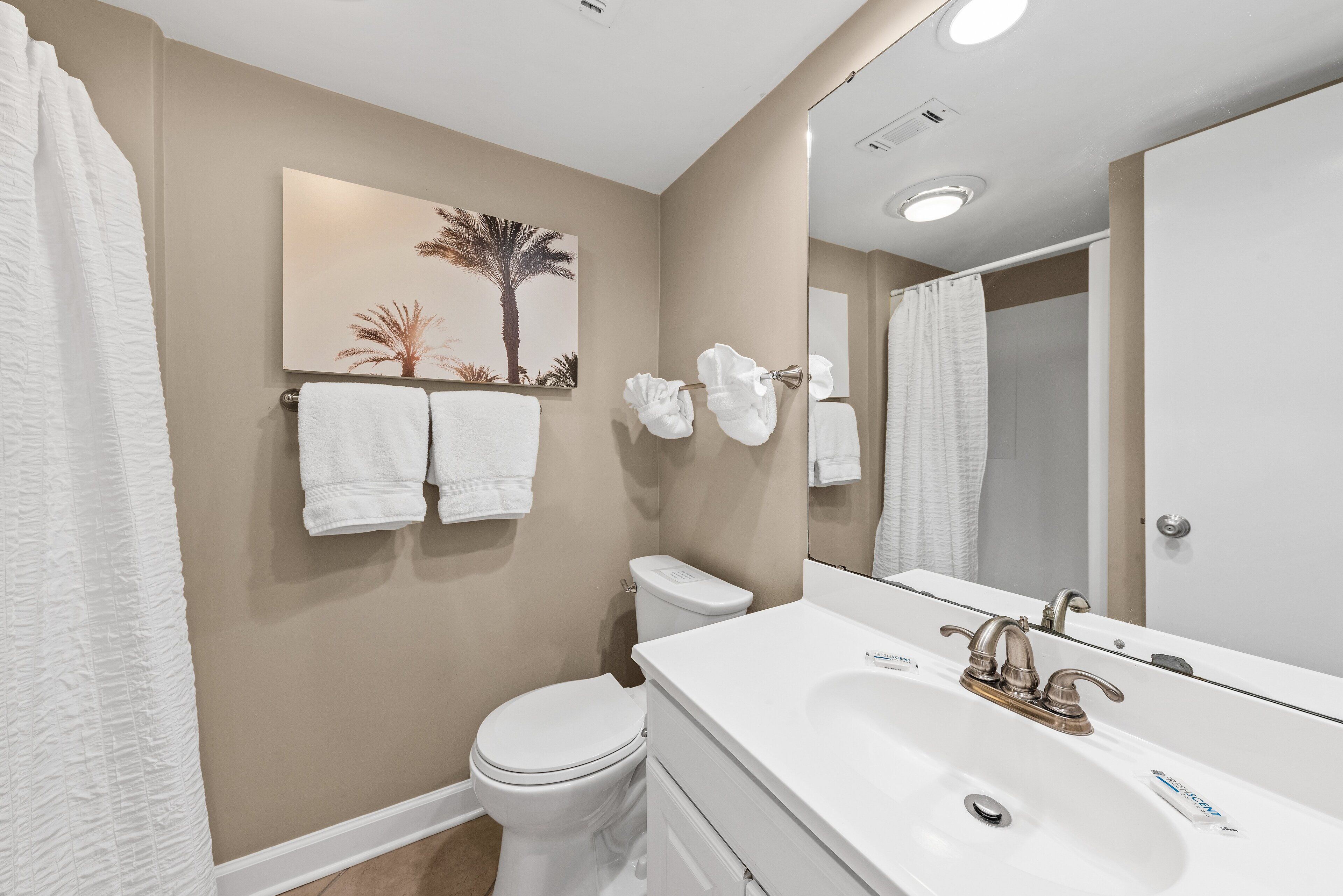 A bathroom with a toilet, sink, and a large mirror. There are towels hanging on the wall and a painting of palm trees.