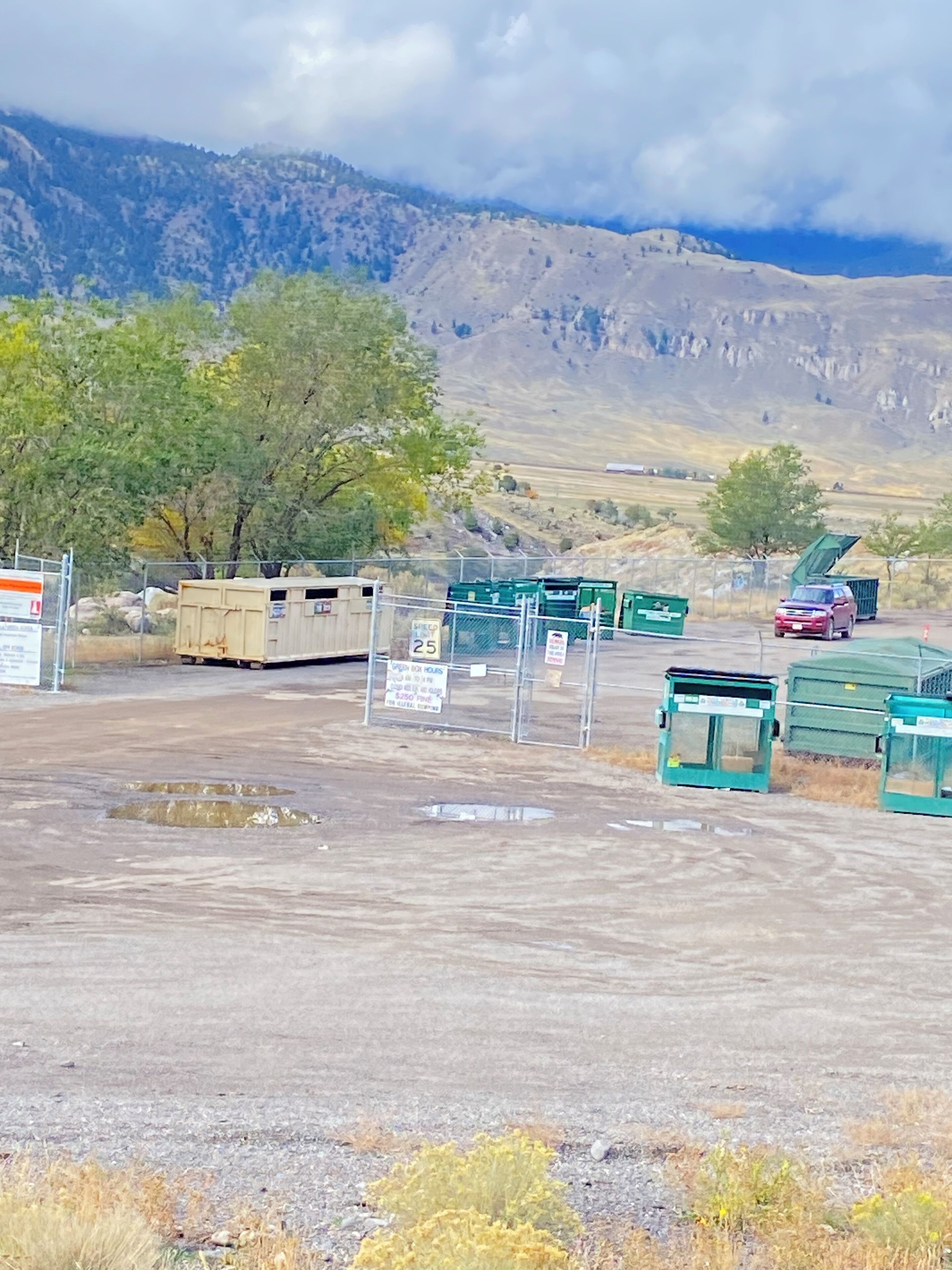 Un parcheggio con cassonetti verdi, un'area recintata e montagne sullo sfondo.