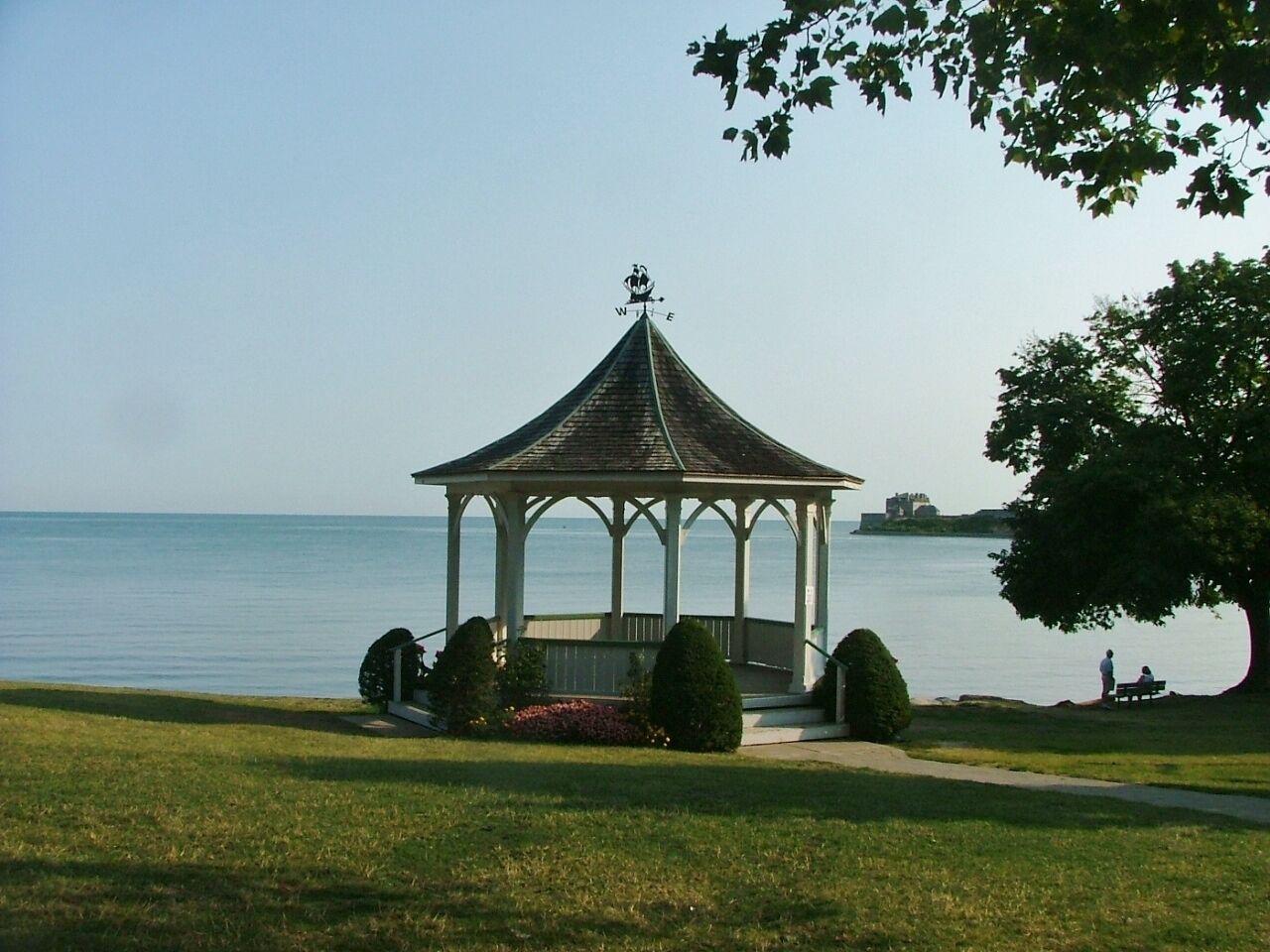 A gazebo with a weather vane, surrounded by grass and a tree, near a body of water.