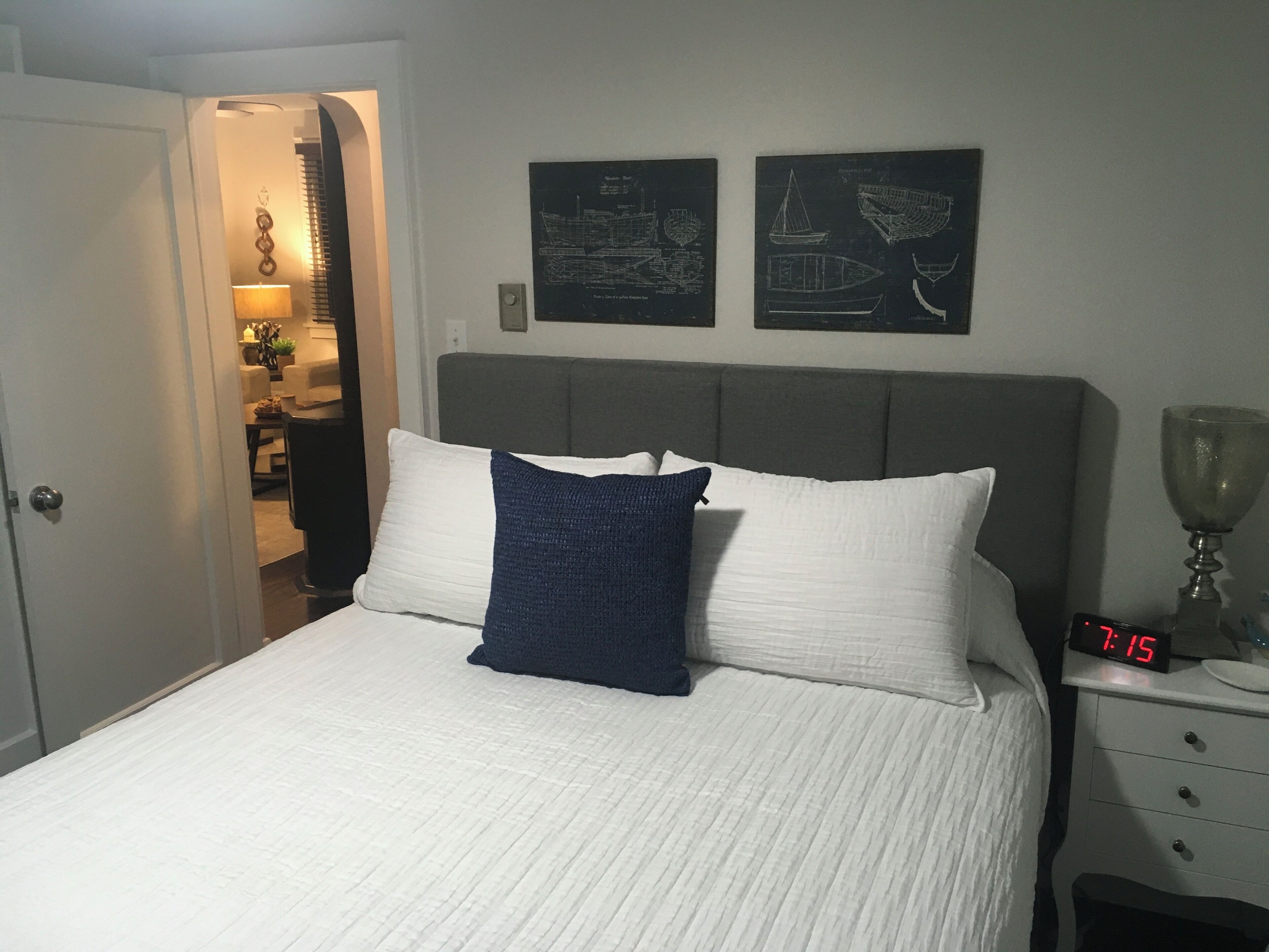 Una cama bien hecha con ropa blanca y almohada azul, una mesita de noche con un reloj, y dos mapas náuticos enmarcados en la pared.