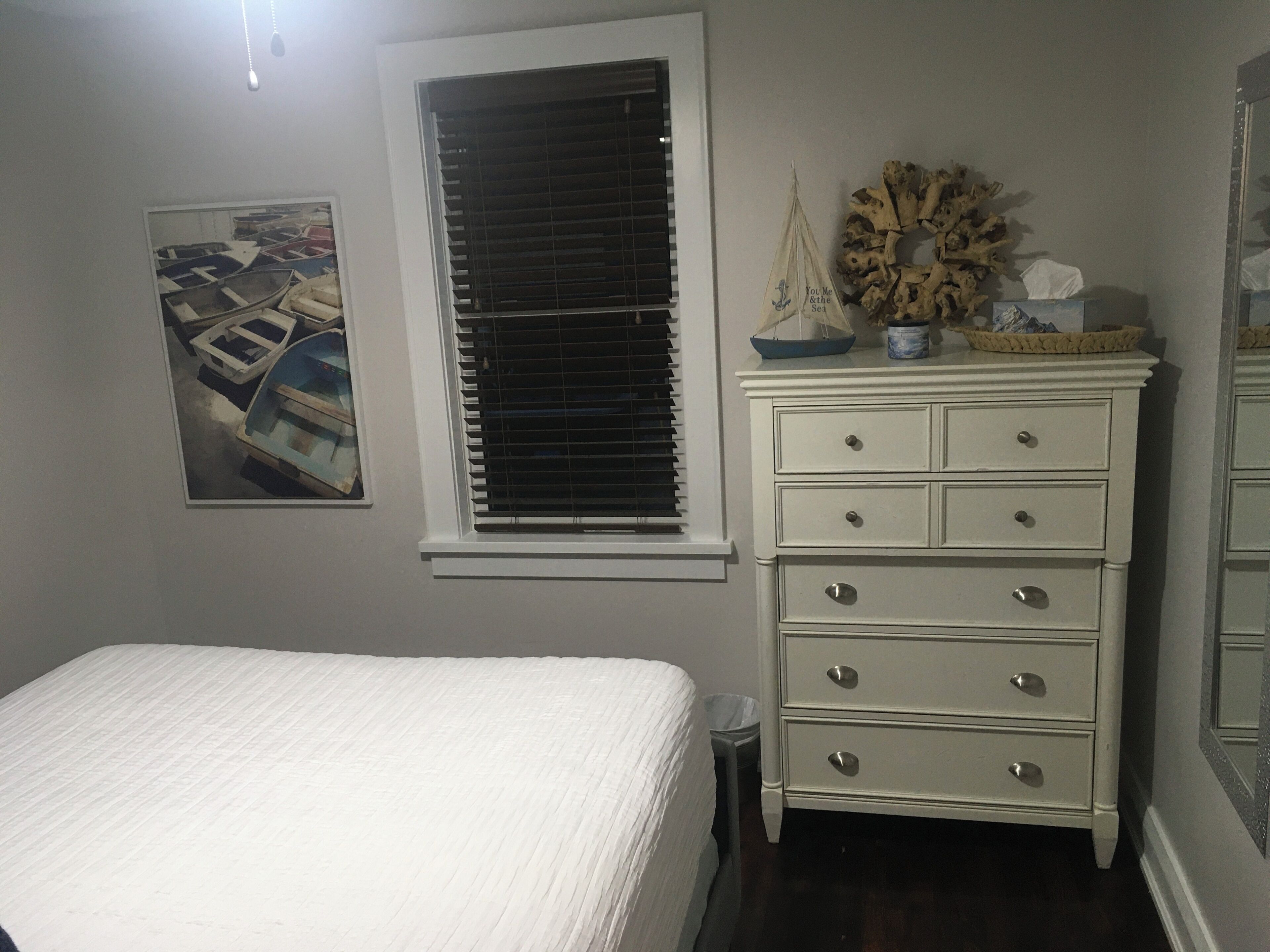 A bedroom with a bed, a dresser, a window with blinds, a wall-mounted picture, and a small trash bin.