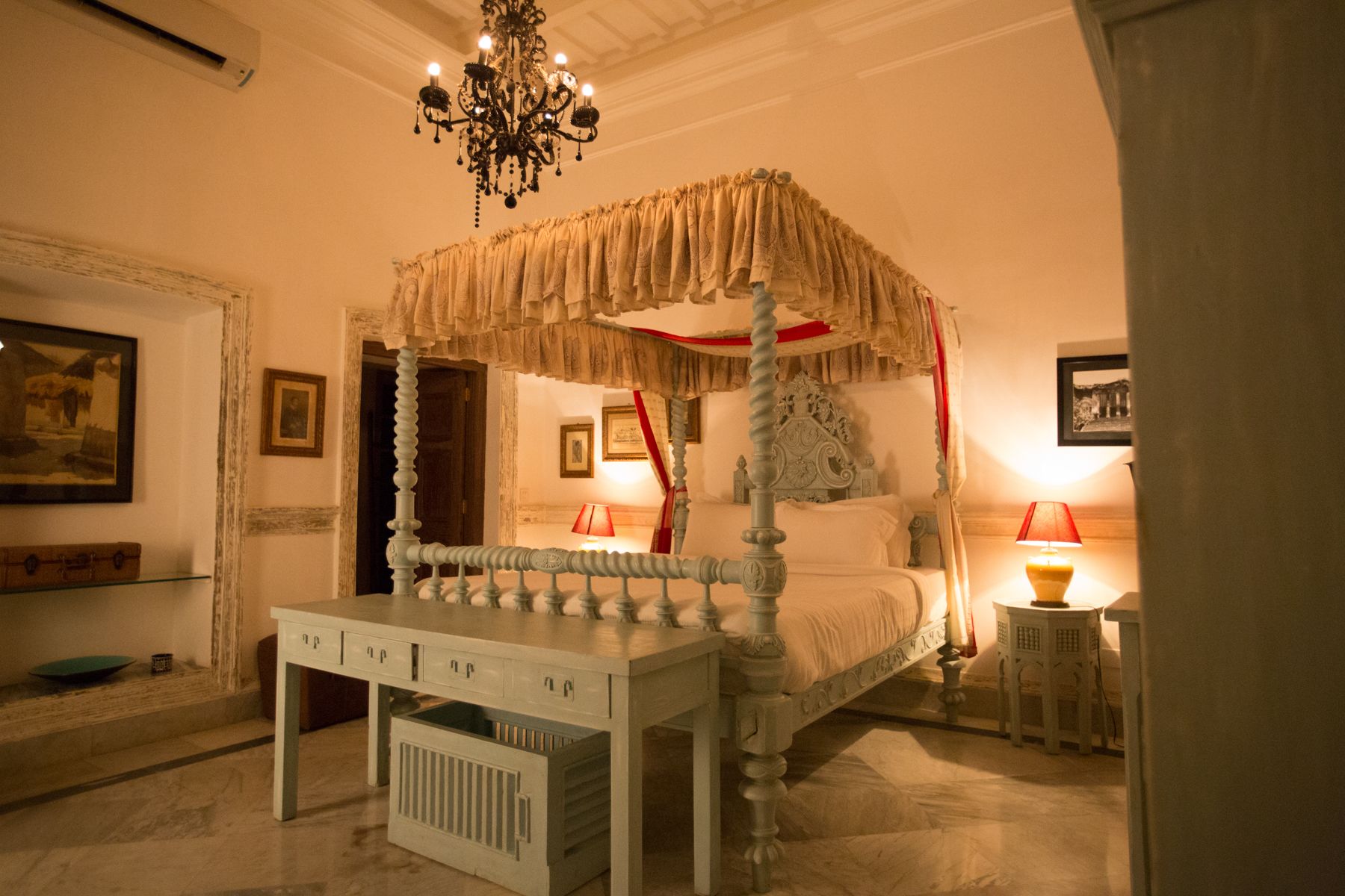 A four-poster canopy bed with a chest of drawers in front, a chandelier, and a bedside table with a lamp.