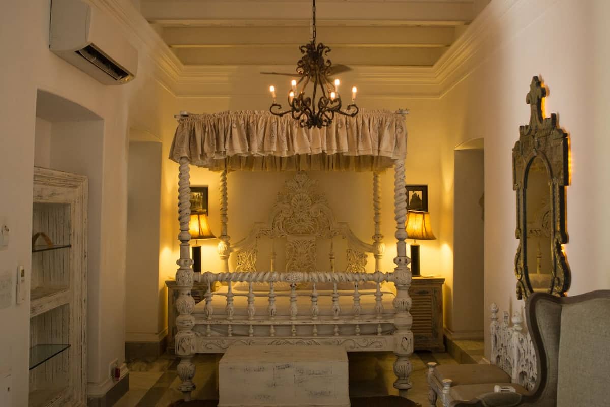 A bedroom with a four-poster bed, a chandelier, a mirror, and a lamp.