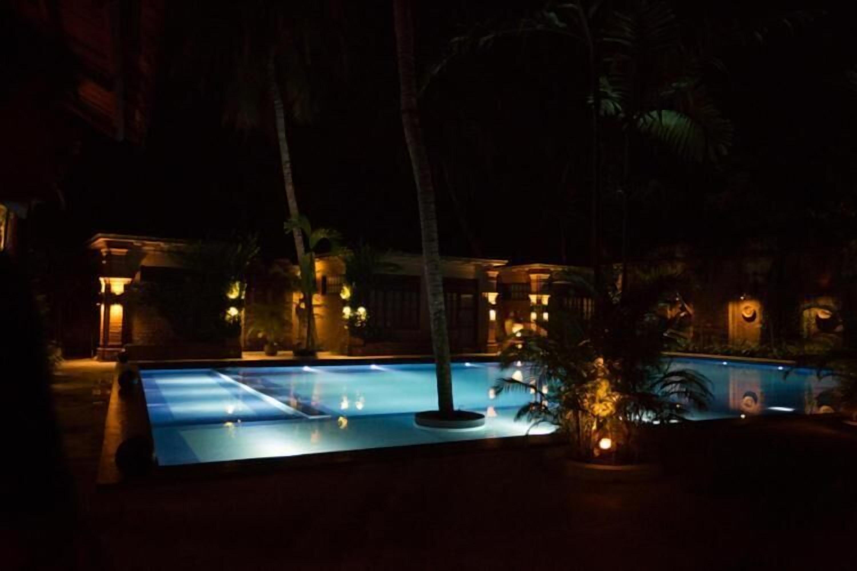A nighttime view of a swimming pool with surrounding palm trees and a building with illuminated windows.