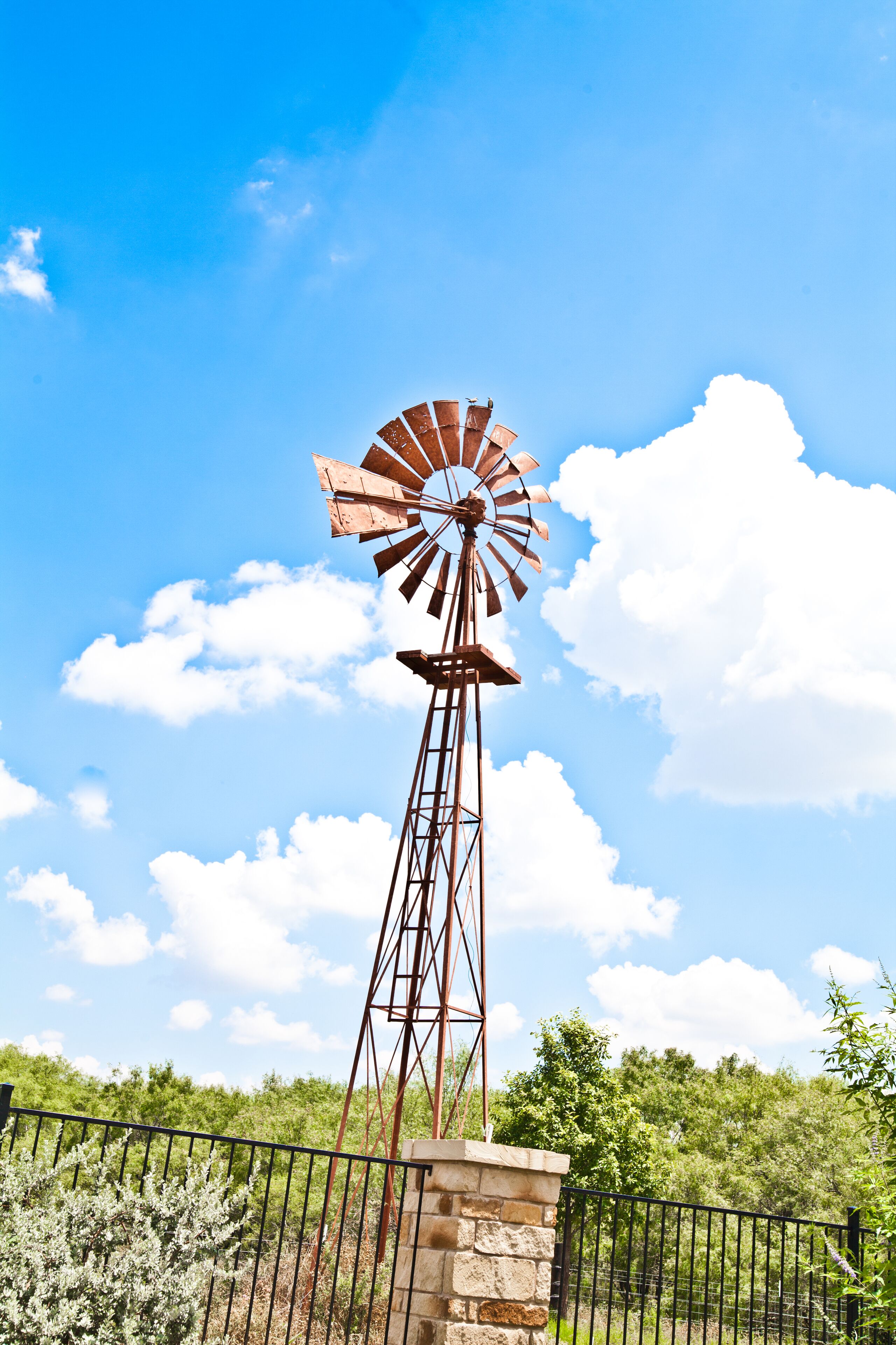 Eine Windmühle mit Metallkonstruktion und steinernen Fundament, umgeben von einem schwarzen Metallzaun und Pflanzen.