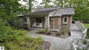 A two-story house with a stone foundation, a wooden deck, and a covered porch with two chairs. - Homestead Hideaway away from it all (Glen Arbor)