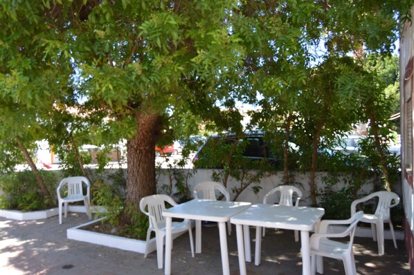 Un área de estar al aire libre con mesas y sillas de plástico blanco, rodeada de follaje y un árbol.