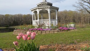 A gazebo with a white structure and a roof, surrounded by a garden with pink tulips and various flowers, set against a backdrop of trees and a body of water. - Scenic Lakeside Chateau with Heated Pool, Hot Tub & Outdoor Kitchen-TV-Fireplace (Belle)