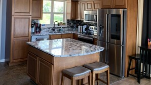 A modern kitchen with wooden cabinets, a large island with stools, stainless steel appliances, and a pendant light. - Scenic Lakeside Chateau with Heated Pool, Hot Tub & Outdoor Kitchen-TV-Fireplace (Belle)