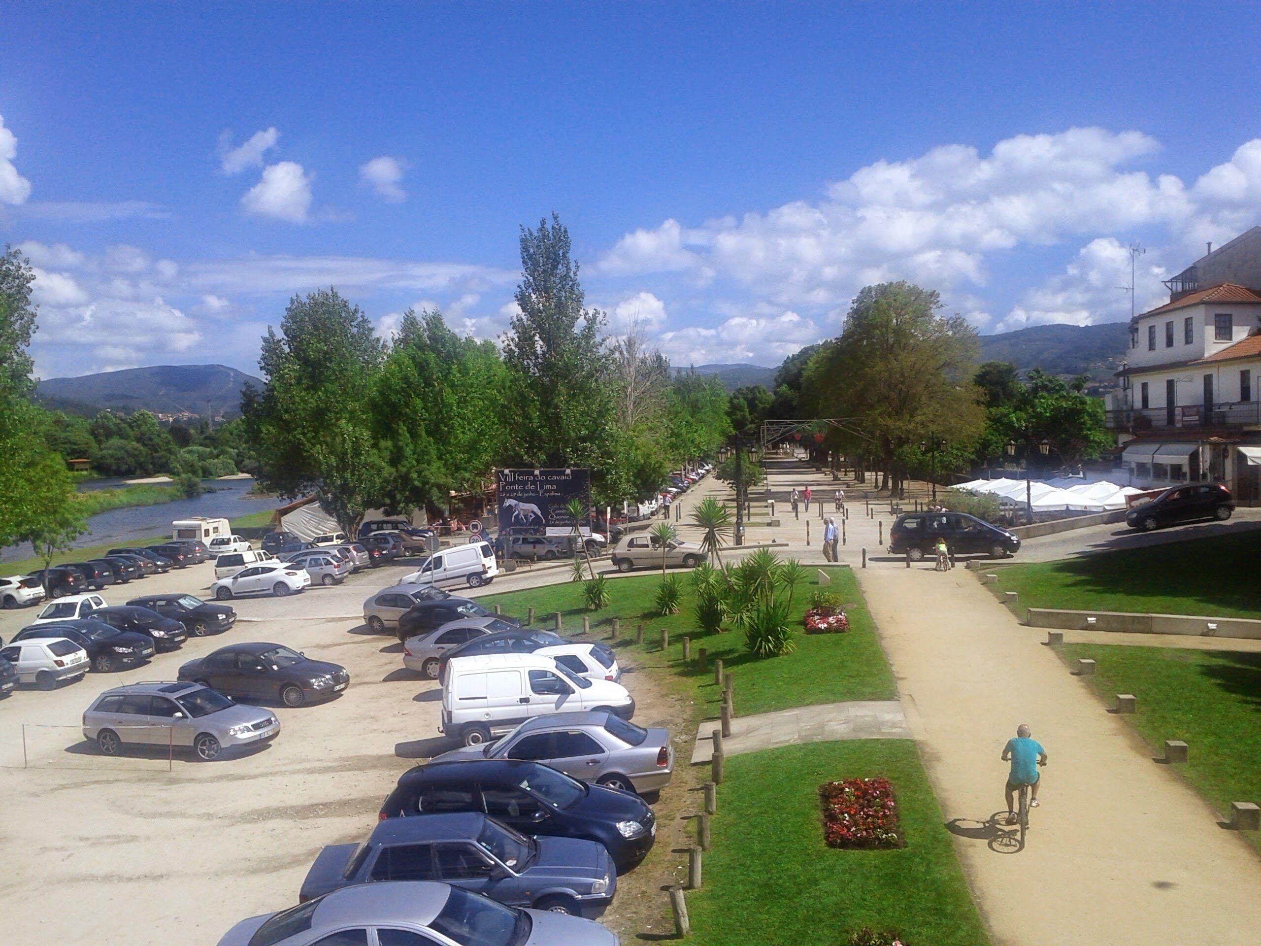A parking lot with several cars and a white van. There is a signboard and a pathway leading to a building.