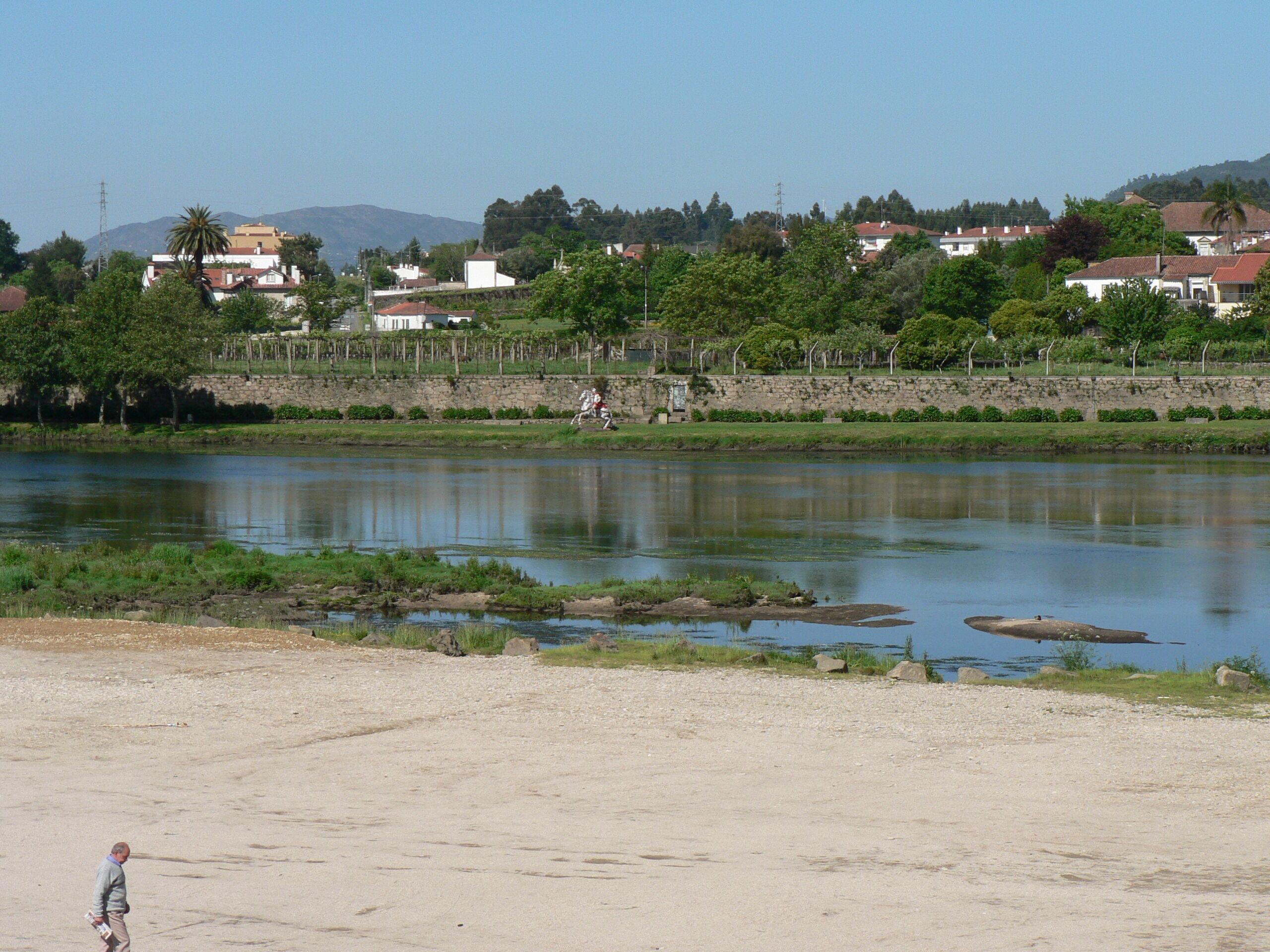 A river with a sandy bank and a few trees. In the background, there are houses and a stone wall.