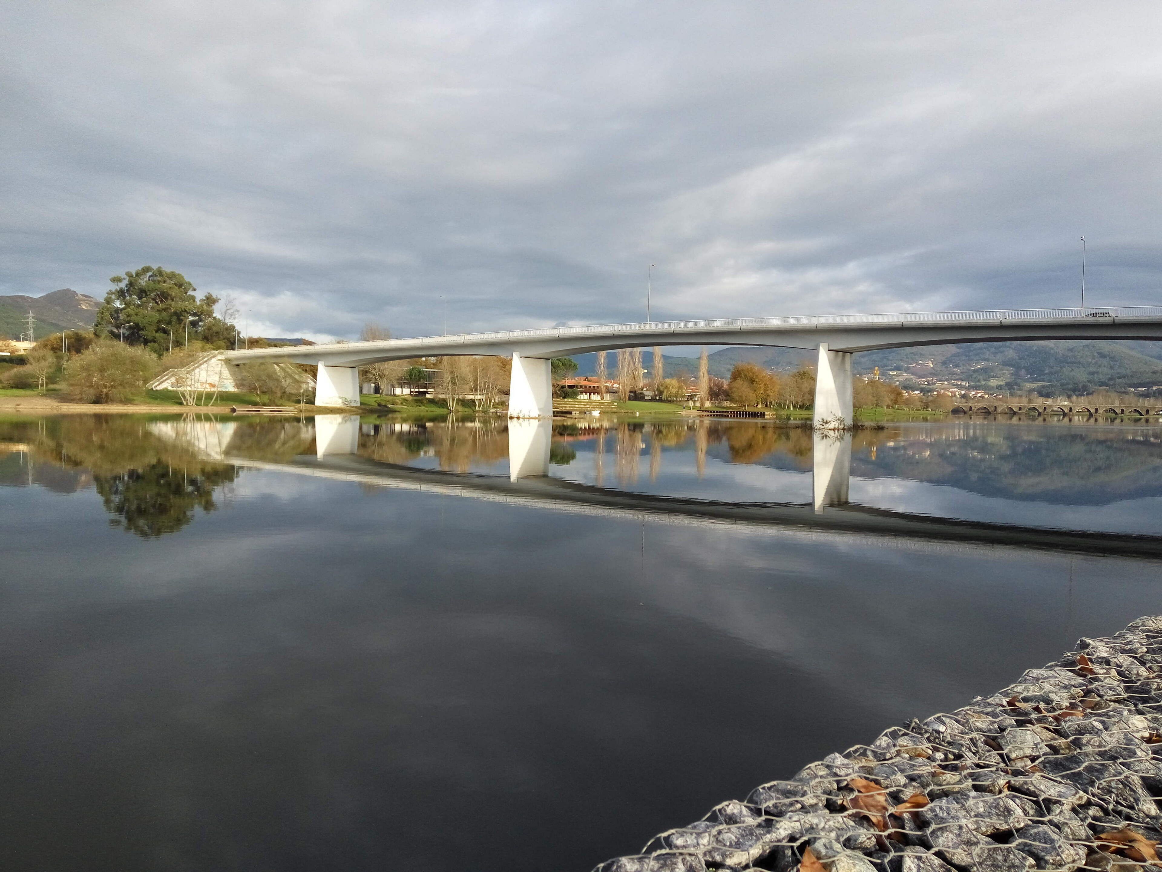 Un pont enjambant une étendue d’eau calme dont le reflet est limpide, entouré de verdure et de bâtiments éloignés.