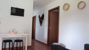 Una sala de estar con un televisor montado en la pared, una mesita con dos sillas y una puerta que da a otra habitación.