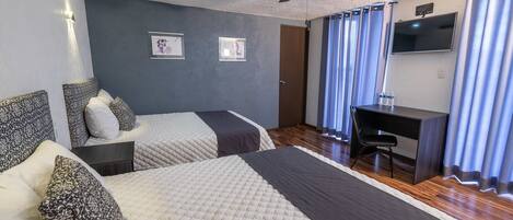 Habitación de hotel con dos camas, un escritorio, una silla, un ventilador de techo y un televisor en la pared.