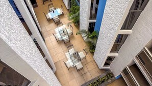 Un patio al aire libre con varios niveles, mesas y sillas, rodeado de edificios con fachadas azules y blancas.