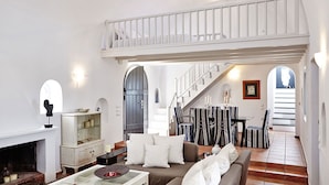 Un salón amplio con techo alto, una chimenea, un comedor con sillas a rayas, y una escalera que conduce a un nivel superior.