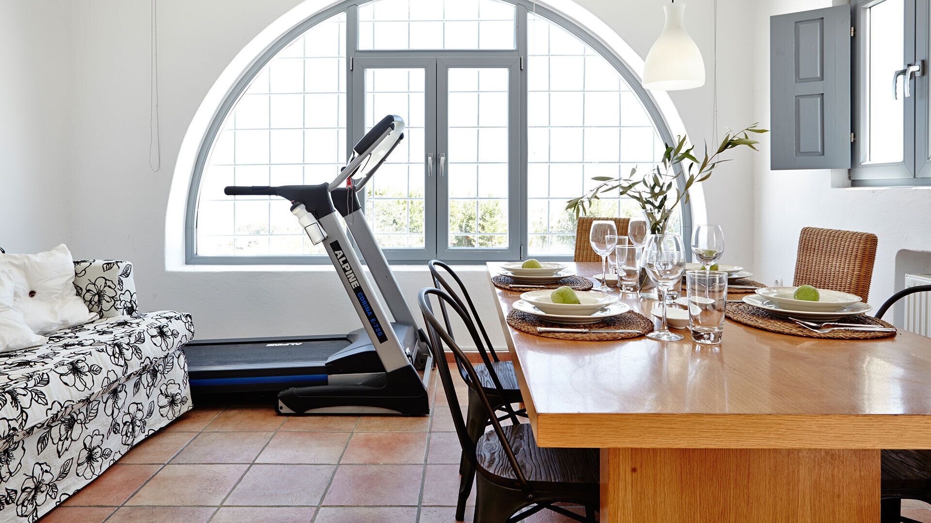 Uma área de refeições com mesa posta para quatro pessoas, uma esteira no canto e uma grande janela em arco que oferece iluminação natural.