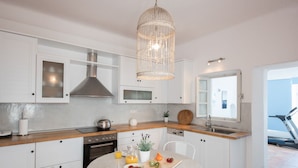 Una cocina moderna con armarios blancos, campana extractora de acero inoxidable, lámpara colgante, mesa de comedor con sillas y vista al mar a través de una ventana.
