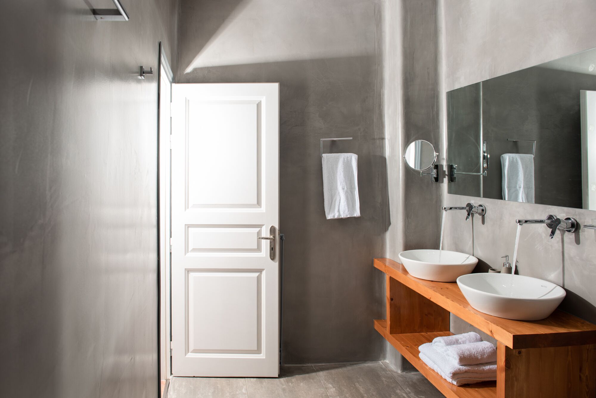 A modern bathroom with a white door, a large mirror, two sinks, and a towel rack.